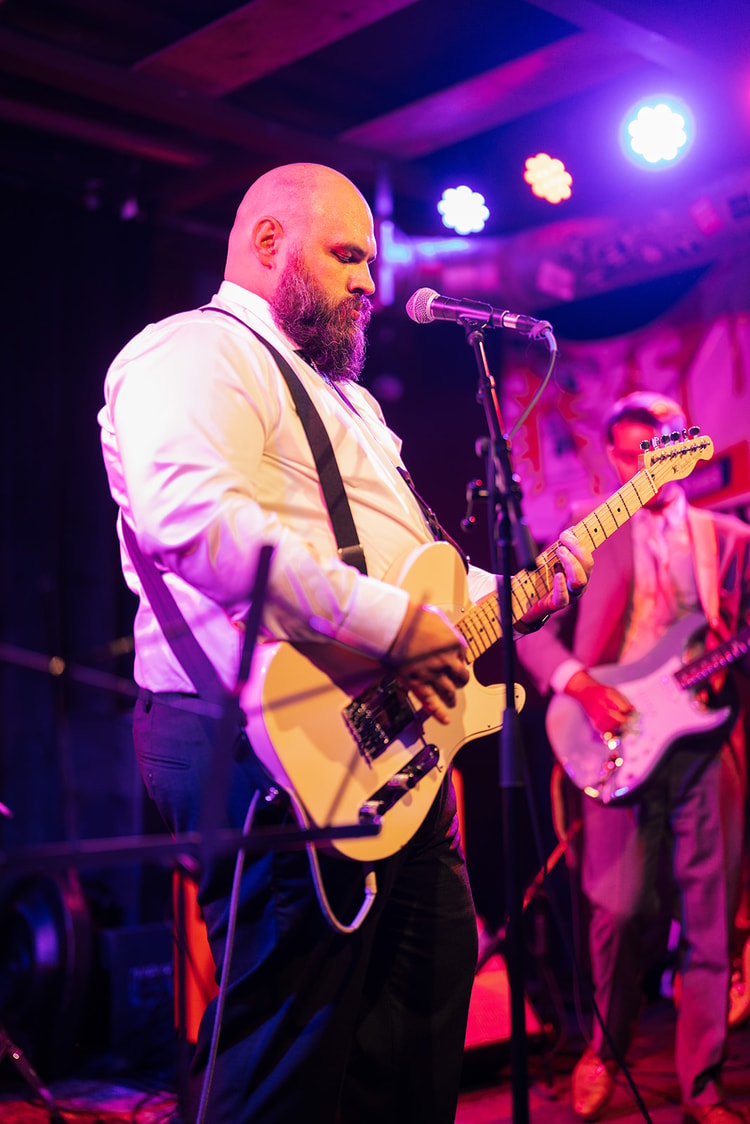 Groom in a white shirt and suspenders playing electric guitar on stage under vivid purple and orange stage lights at the reception