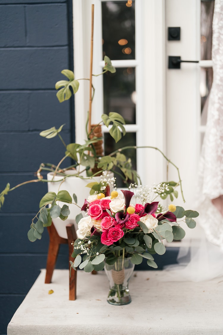 Wedding bouquet of hot-pink roses, deep burgundy calla lilies, white blooms, and eucalyptus in a glass vase on a white ledge beside a navy blue building, with the lace wedding gown hanging at the edge of frame