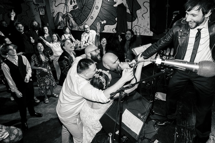 Black and white reception moment: bride and groom leaning toward the vocalist's microphone on stage while guests cheer and raise their hands in the crowd behind them